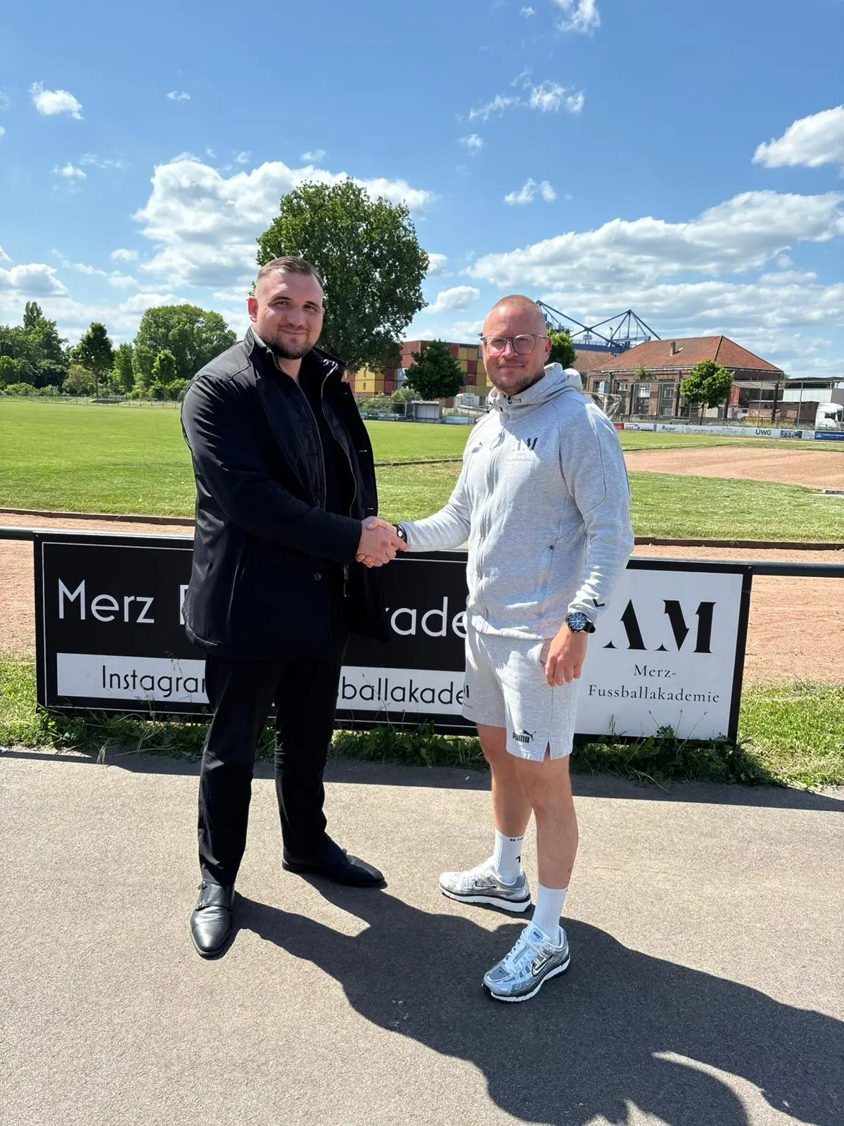 Zwei Personen geben sich am Sportplatz die Hand, im Hintergrund eine Bande der MERZ Fußballakademie